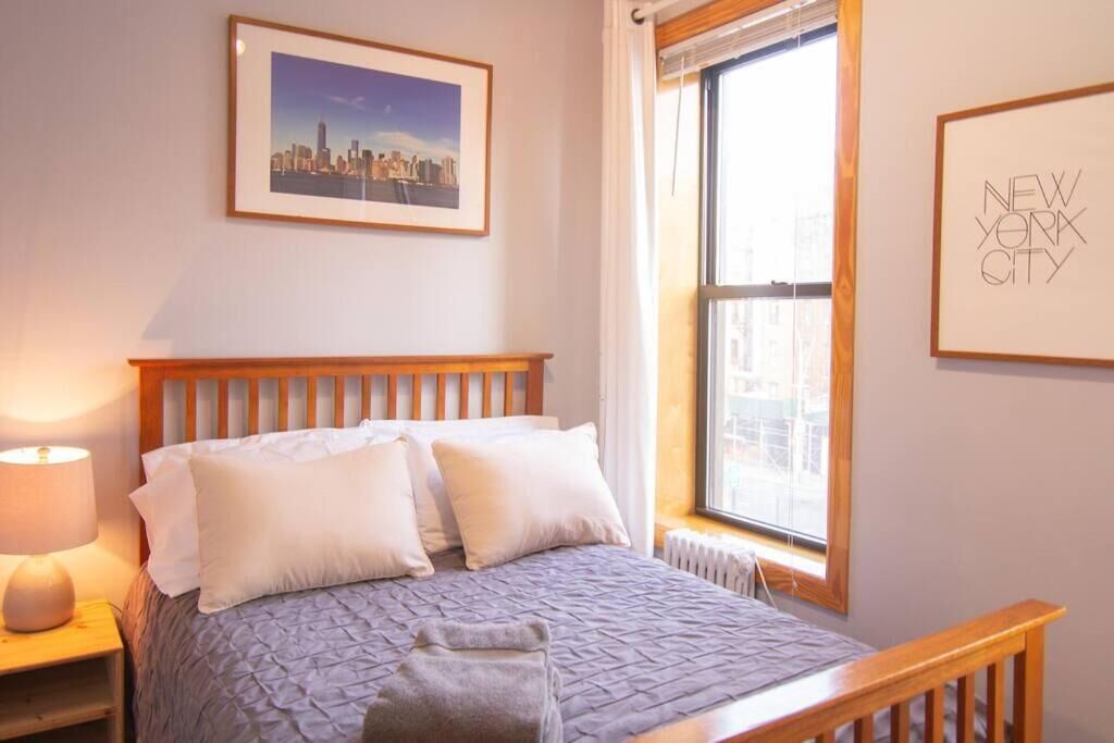 Una cama bien hecha con cabecera, una lámpara de noche, un cuadro enmarco con una vista urbana y una ventana que ofrece vistas a edificios.