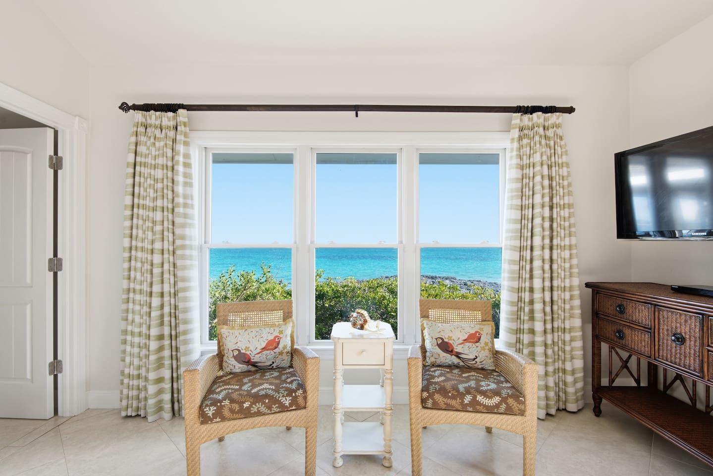 Une pièce avec vue sur l’océan, deux fauteuils en osier avec des coussins, une petite table blanche et un buffet en bois.