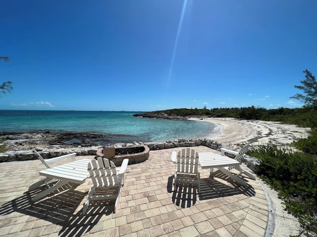 Eine Terrasse direkt am Strand mit Adirondack-Stühlen und einem Feuerkorb, mit Blick auf das klare, blaue Meer und eine felsige Küstenlinie.