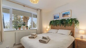 Quarto com cama, luminárias de cabeceira, janela com cortinas, radiador e planta suspensa na parede.