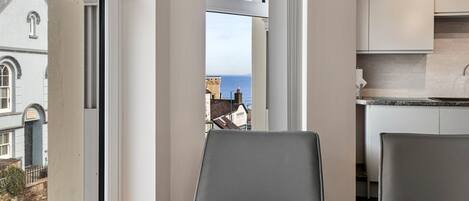 Un coin repas avec une table dressée pour deux personnes, une vue sur la mer à travers de grandes fenêtres et, au fond, une cuisine moderne.