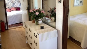 Un dormitorio con cama, cómoda con cajones, espejo, un jarrón con flores e impresión botánica enmarcada en la pared.