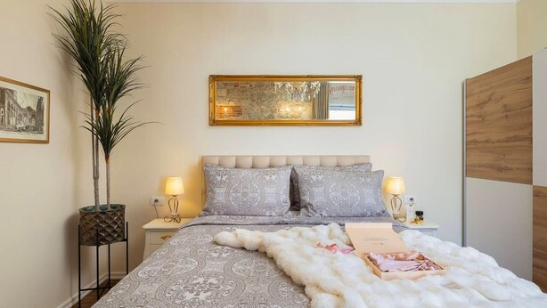 A bedroom with a bed, two bedside tables, a nightstand, a large mirror, a chandelier, and a potted plant. - Modern Comfort in Heart of Split (Split)