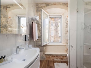 Ein Badezimmer mit Steinwand, Fenster, Waschbecken mit Pflegeprodukten und Duschbereich.