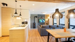 A modern kitchen with wooden cabinets, a central island, pendant lighting, a sofa, and a dining table with a vase. - Apartments pri fencu (Jesenice)