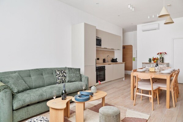 En moderne stue med en grøn sofa, et spisebord dækket til fire personer, og et køkken med hvide skabe og en mikrobølgeovn.