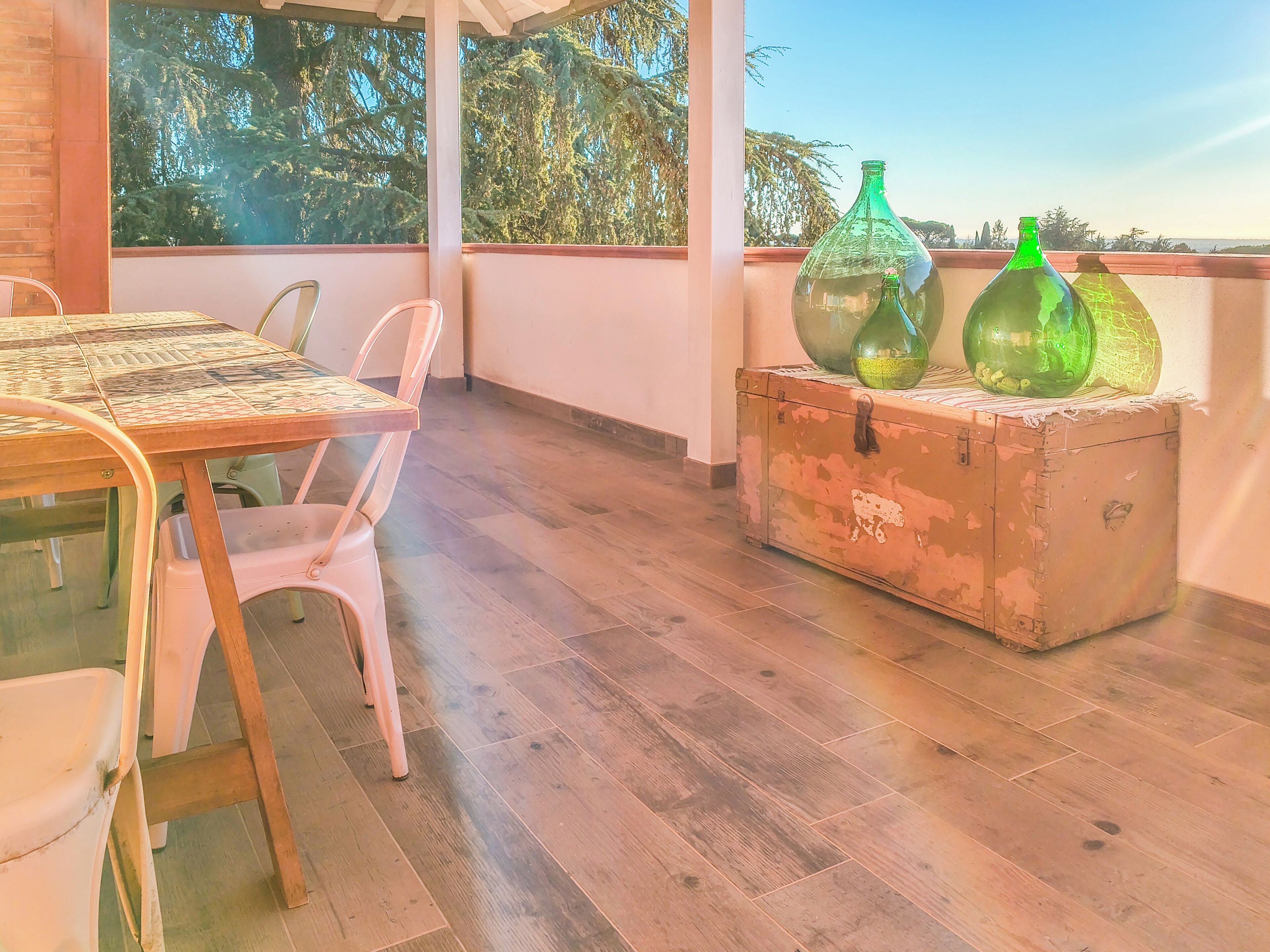 A wooden table with chairs, a vintage trunk, and green glass bottles on a balcony with a view of trees.