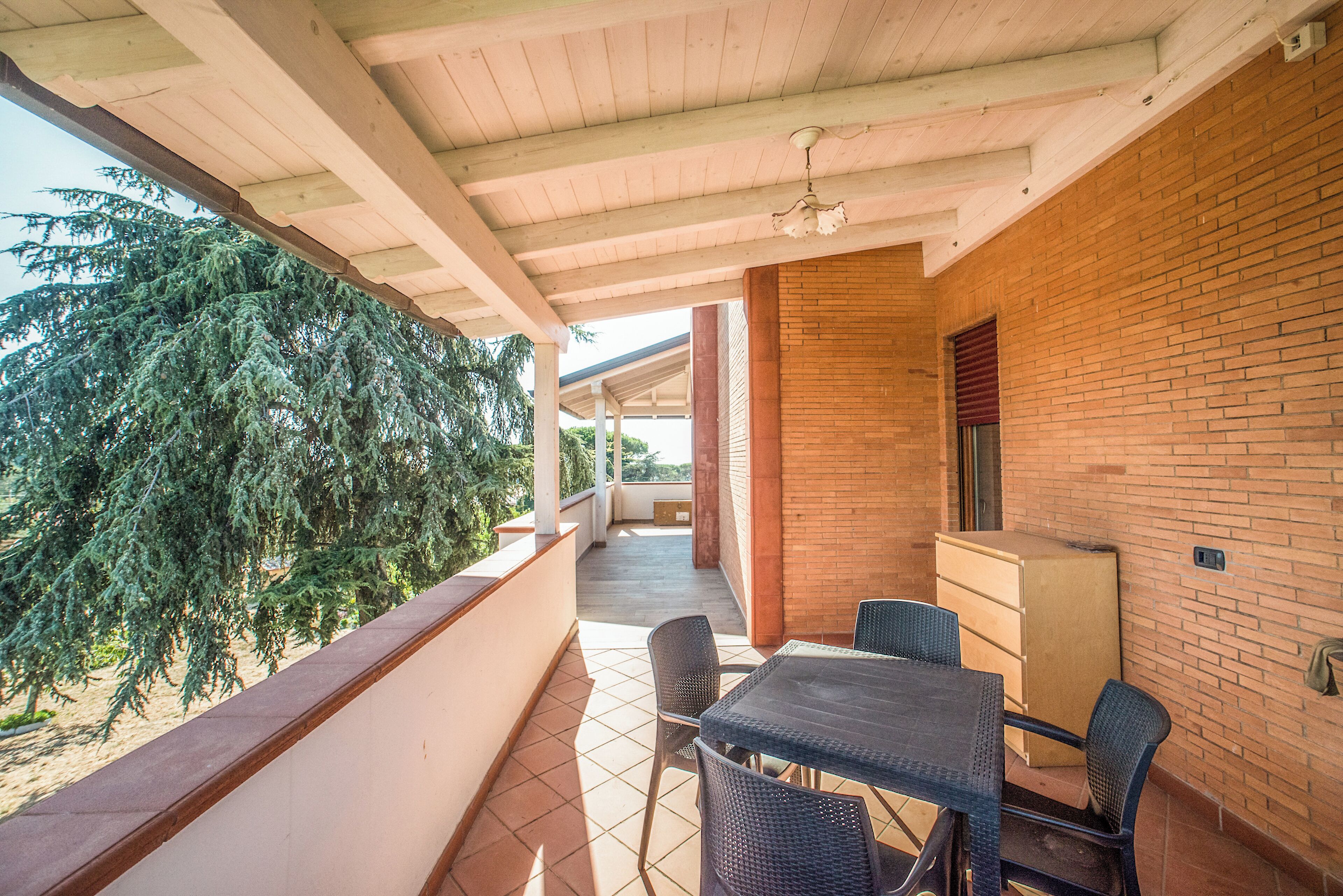 A covered balcony with a table and chairs, a brick wall, and a view of trees.