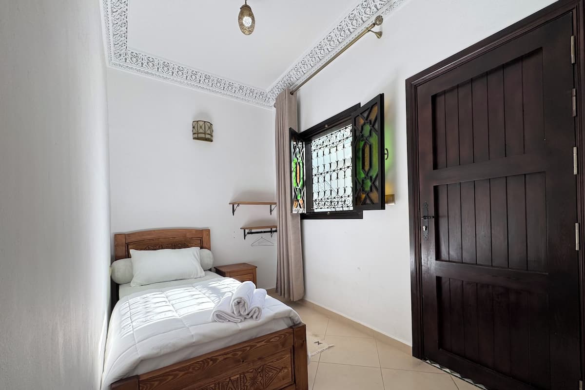 Un dormitorio con una cama de madera, una puerta oscura, una ventana con celosías decorativas y una lámpara fijada a la pared.