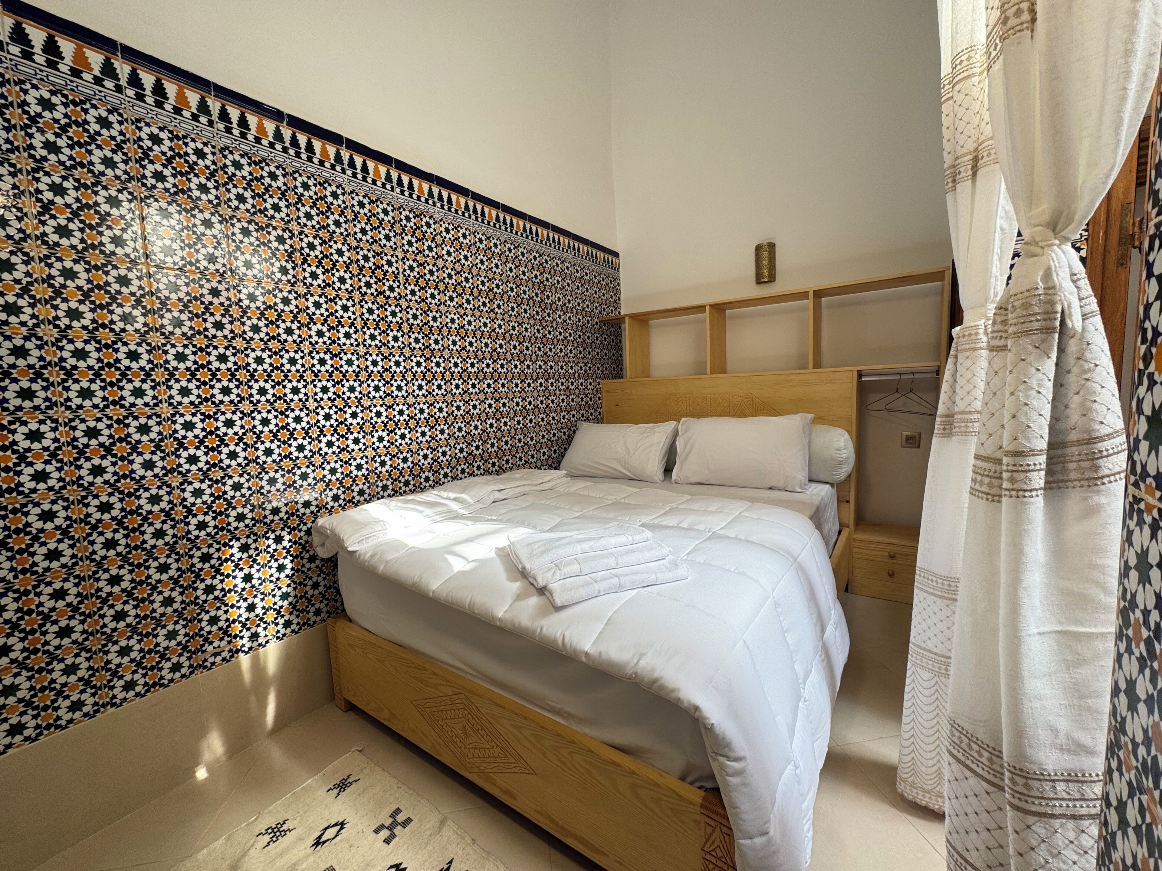 Un dormitorio con pared de baldosas estampadas, cabecera de madera, cama con ropa de cama blanca y cortina colgante.