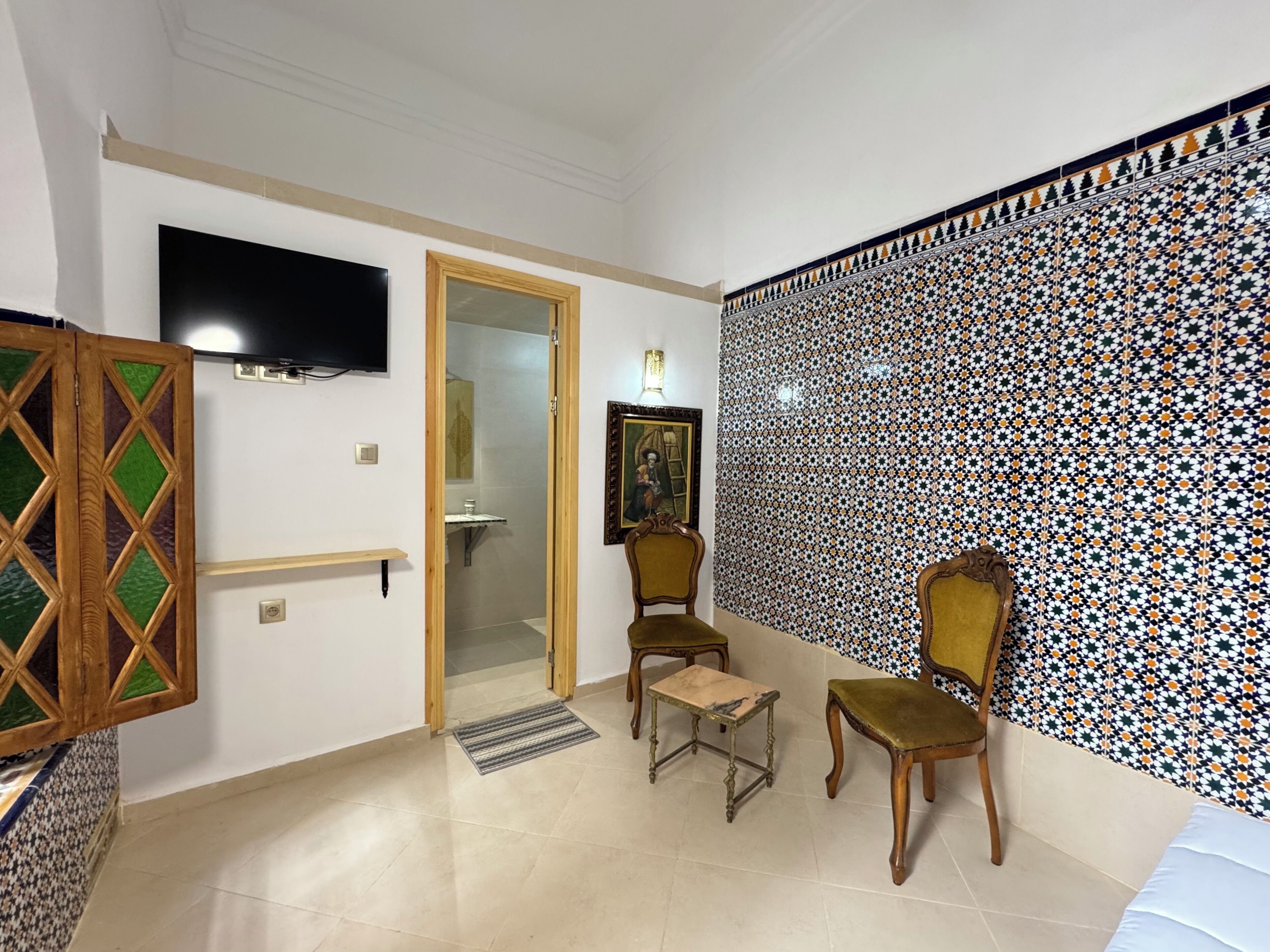 Una habitación con televisión, una puerta que da al baño, dos sillas, una mesita y un papel tapiz estampado.