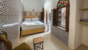 A bedroom with a bed, a small table, a hanging light, a window with decorative ironwork, and patterned walls.