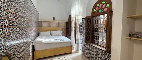 A bedroom with a bed, a small table, a hanging light, a window with decorative ironwork, and patterned walls.