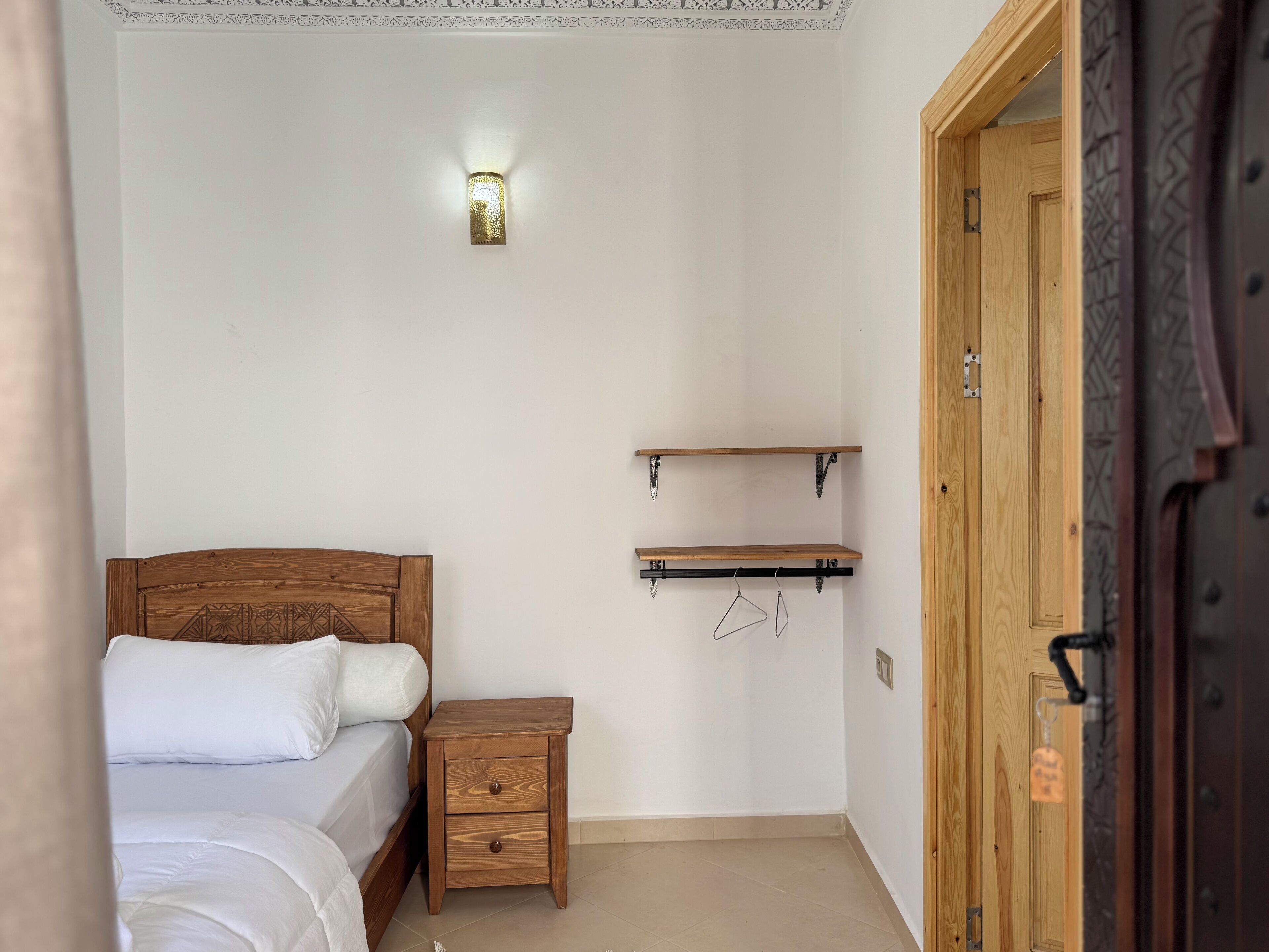 A bedroom with a wooden bed, a nightstand, a wall-mounted shelf, and a door.