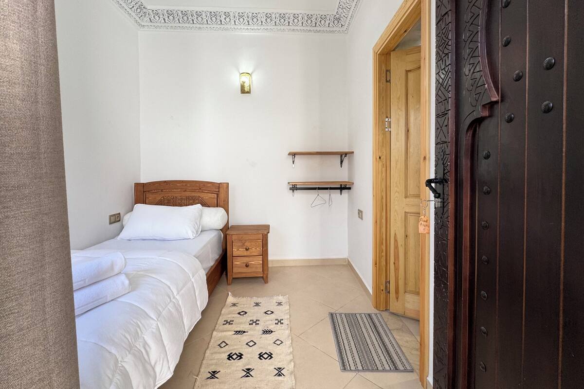 Un dormitorio con cama, mesita de noche, lámpara colgante, puerta de madera y una alfombra estampada.