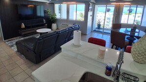 Un soggiorno moderno con un divano in pelle nera, un televisore a schermo piatto, una zona pranzo con un tavolo e sedie bianche e ampie finestre con vista sull'esterno.