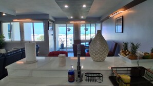 A modern kitchen with a large island, a dining area with a red chair, and a view of the outdoors through large windows. - NEWLY RENOVATED, Tranquil oceanview gated community. Close to Beach and Airport (Río Grande)