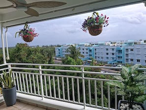 A balcony with hanging flower baskets, a potted plant, and a view of a blue building complex. - NEWLY RENOVATED, Tranquil oceanview gated community. Close to Beach and Airport (Río Grande)