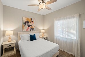 A bedroom with a bed, two bedside tables, a ceiling fan, a window with sheer curtains, and a painting on the wall. - Desert Sky Escape in Marana (Marana)