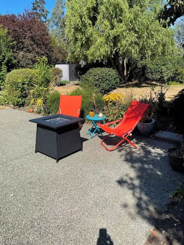 An outdoor seating area with a black table and two red chairs, surrounded by greenery and a tree. - Swan Lake Oasis (Victoria)