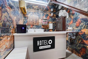 A hotel reception desk with a marble wall, a sign indicating 'HOTELO Company Serviced', and a small fan above the counter. - Hotel O Isbt Sector 43 Bus Terminal (Chandigarh)