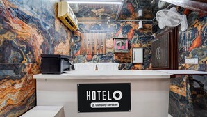 A hotel reception desk with a marble wall, a sign indicating it is company-serviced, a fan, and a small window. - Hotel O Isbt Sector 43 Bus Terminal (Chandigarh)