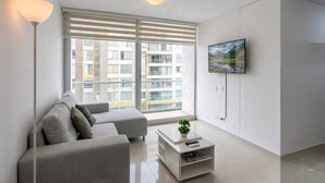 Uma sala de estar moderna com um sofá cinzento, uma mesa de centro branca, um abajur e uma TV fixada na parede.