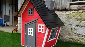 Una casa pequeña de color rojo y gris con techo negro, una puerta y ventanas, ubicada en un área cubierta de césped con un muro de piedra al fondo.