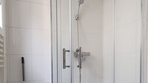 Un espace douche fermé par des parois de verre, doté d’un pommeau de douche à main, d’un chariot de nettoyage bleu avec une serpillière et d’un mur recouvert de carreaux blancs.