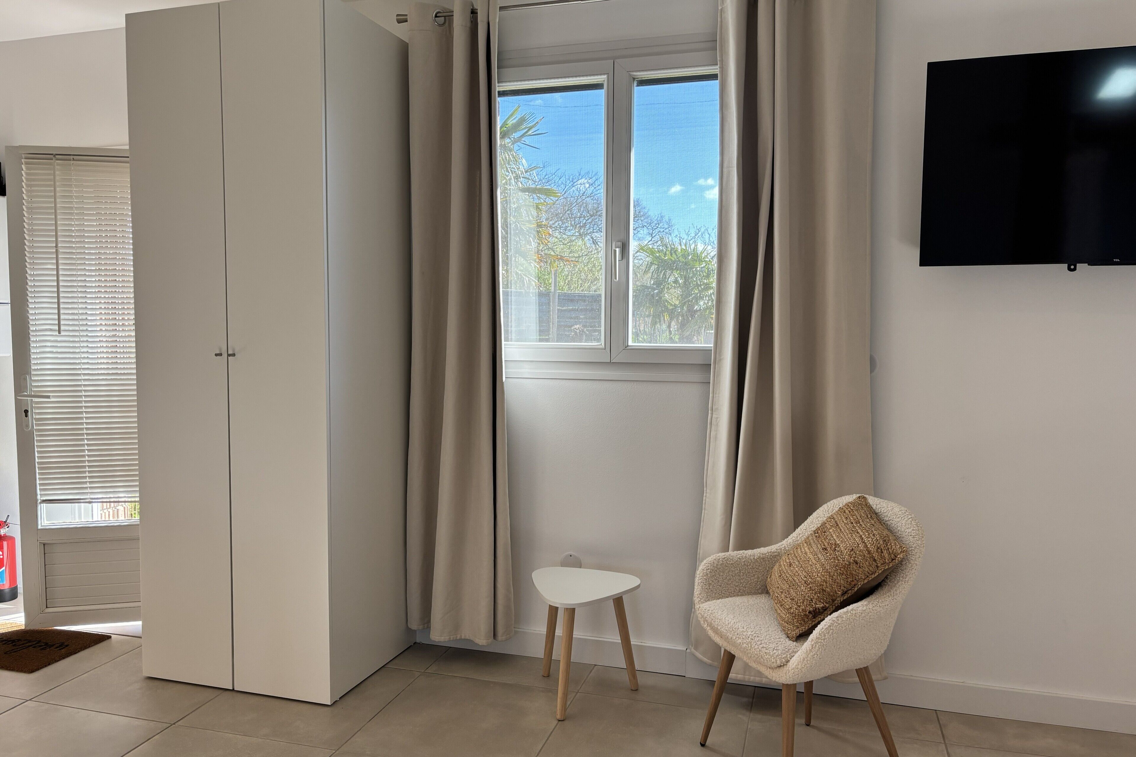 Ein Zimmer mit einer Schiebetür, einem kleinen weißen Tisch, einem gepolsterten Stuhl, einem Wandfernseher und einem fenster mit Vorhängen.