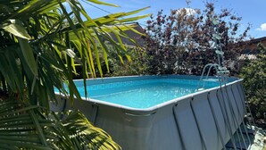 An above-ground swimming pool with a ladder, surrounded by lush greenery and a clear blue sky. - Loft with Private Pool, Jacuzzi, AC & Garden – Peaceful Countryside (Saint-Grégoire)