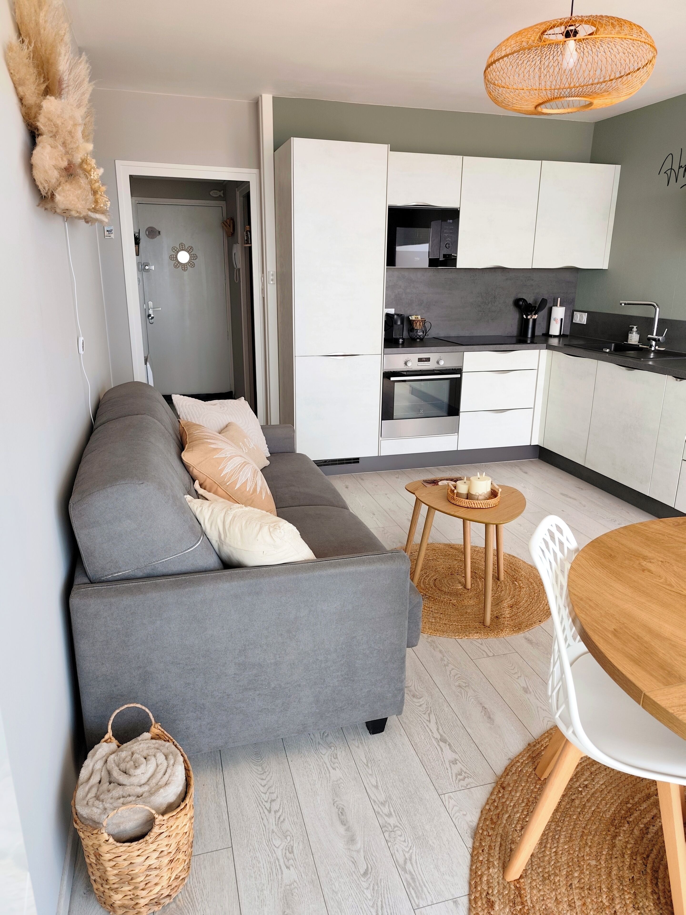 Ein stilvolles Wohnzimmer mit grauem Sofa, einem kleinen Holzesstisch, einer Küchenzeile mit weißen Schränken und einem Korbwäschesammler.