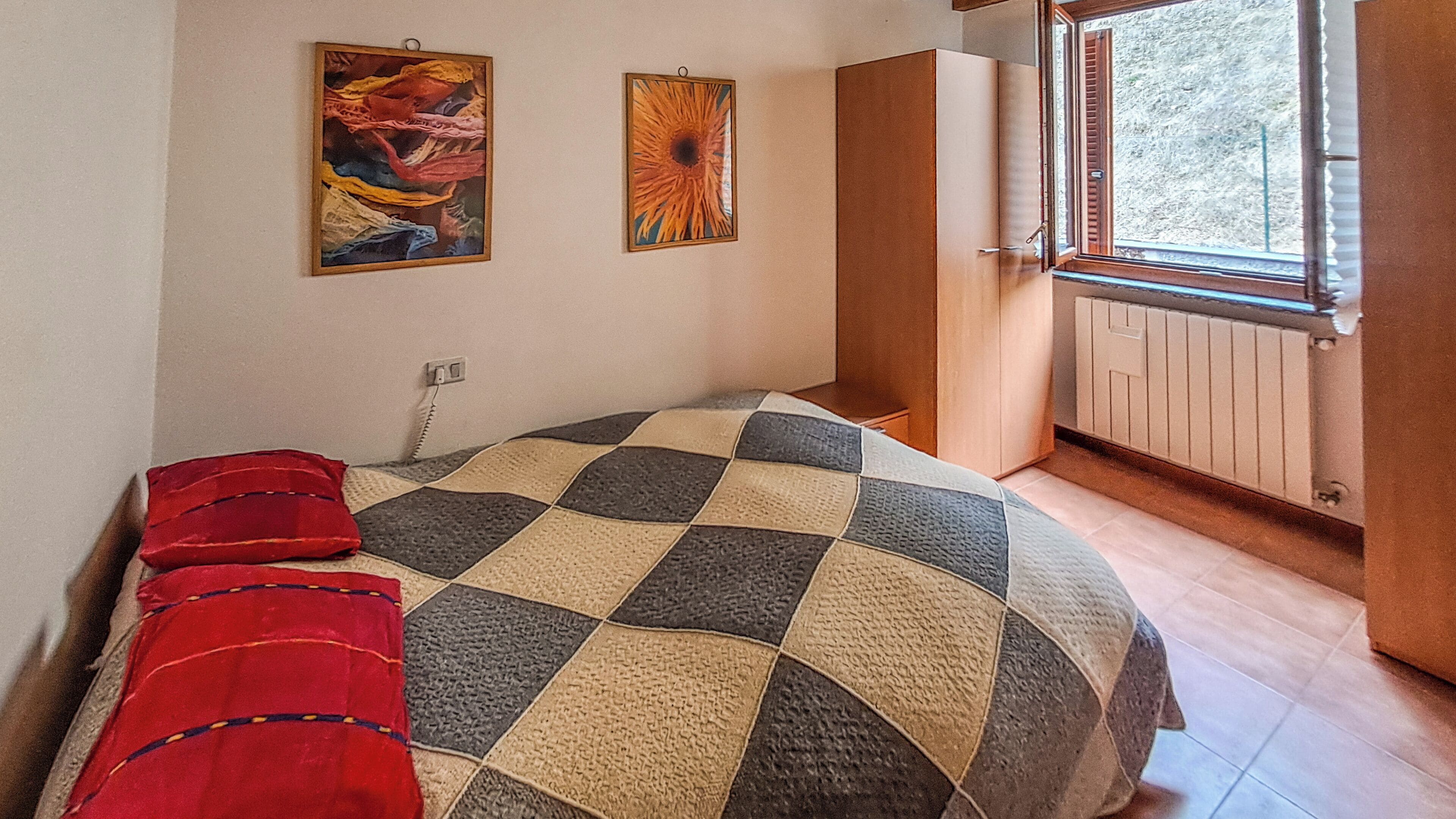 Une chambre avec une couvre-lit à carreaux, deux tableaux accrochés au mur, une fenêtre donnant sur un paysage agréable et une porte menant à l’extérieur.