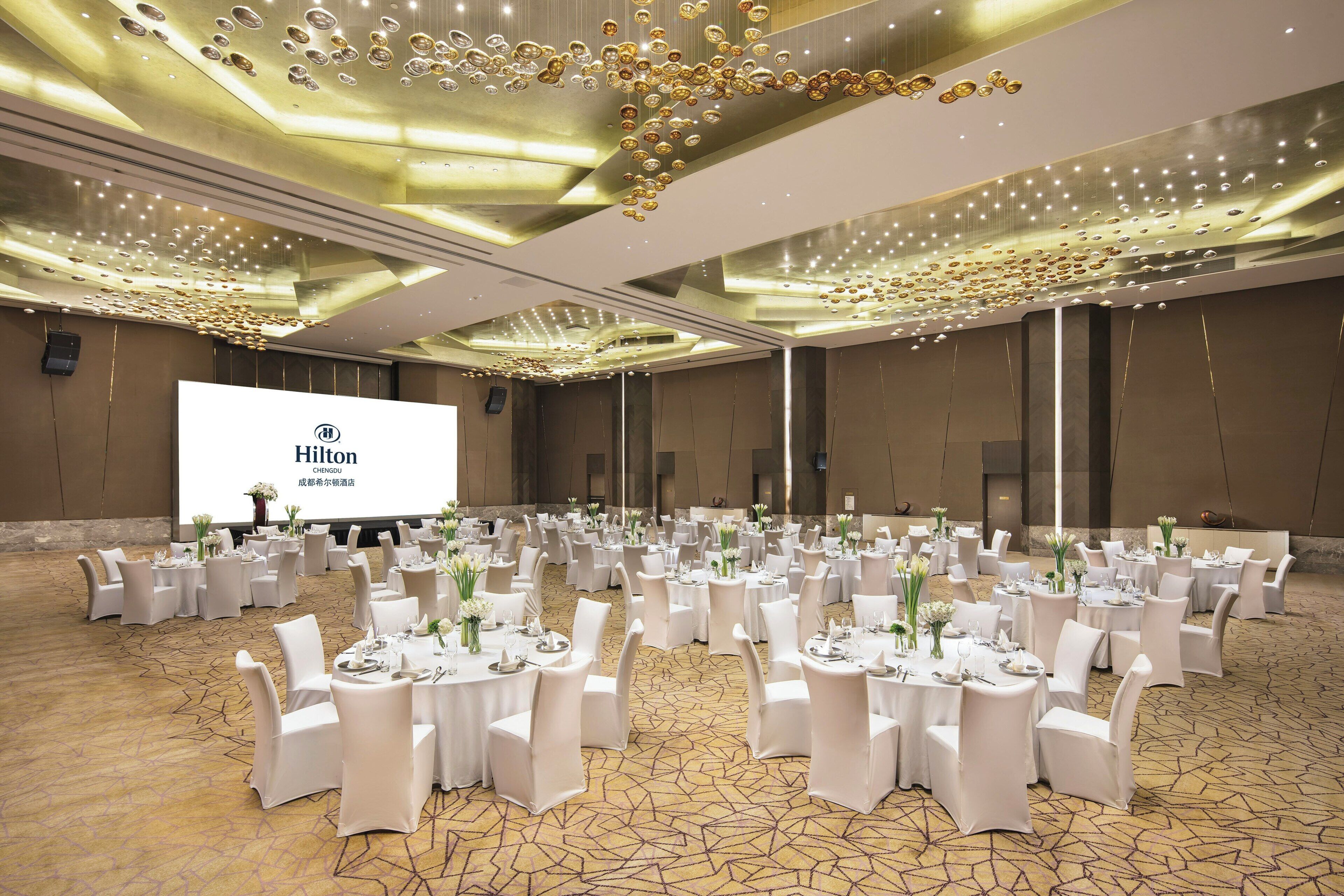 A large, well-lit banquet hall with round tables set for events, a large screen displaying the Hilton logo, and a modern ceiling design.