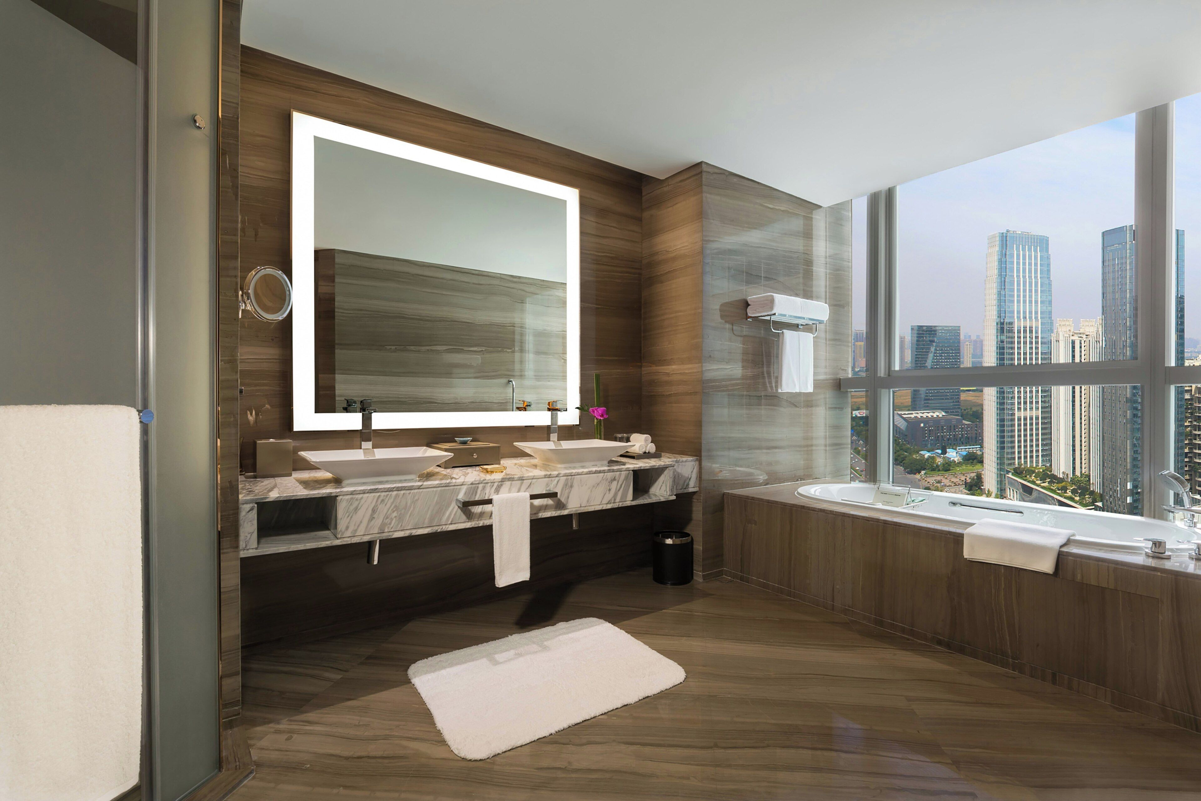 Ein modernes Badezimmer mit einem großen Spiegel, zwei Waschbecken, einer Badewanne und Blick auf eine Stadtlandschaft durch große Fenster.