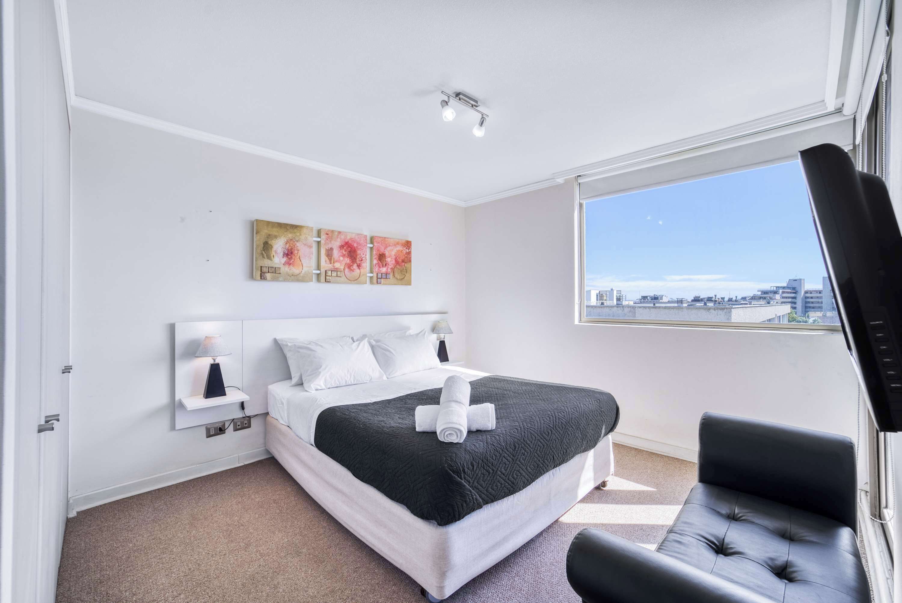 Habitación de hotel moderna con una cama grande, un sillón de cuero negro, vistas a la ciudad y obras de arte en la pared.