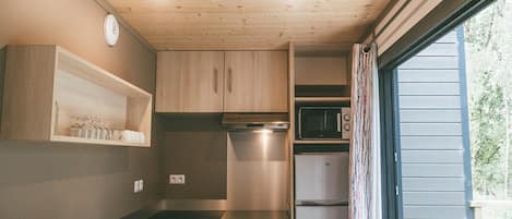 A compact kitchen with wooden cabinets, a microwave, a refrigerator, and a window with curtains.