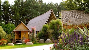 A traditional wooden house with a thatched roof, surrounded by lush greenery and flowering plants. - Villa (Malansac)