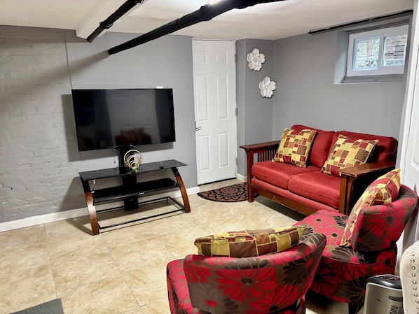 A living room with a flat-screen TV, a red sofa with patterned cushions, a wooden coffee table, and a decorative wall piece.