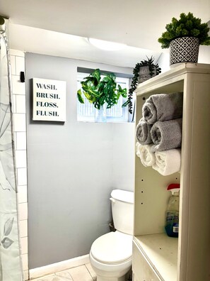 Un baño con inodoro, toallero con toallas, una planta en maceta y un cartel en la pared.