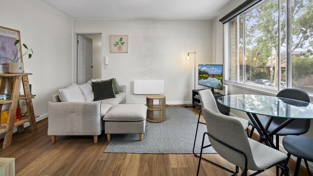 Un salon moderne avec un canapé, un pouf, une table en verre et des fauteuils. On y trouve également un téléviseur, une lampe et une bibliothèque.