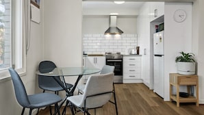 Une cuisine compacte avec des meubles blancs, une hotte en acier inoxydable, un four encastré, un réfrigérateur et un coin repas comprenant une table et des chaises en verre.