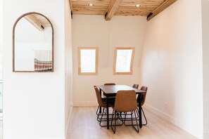 A dining area with a wooden table and chairs, two windows, and a mirror on the wall.