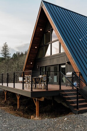 A modern house with a wooden deck, a grill, and a staircase leading to the entrance. - The LuXRae House, Modern A-Frame on 6 Private Acres in the Gorge (Lyle)