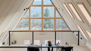 Une pièce dotée d’une grande verrière, de lambris en bois, d’une table avec des chaises et offrant une vue sur les arbres et les montagnes.