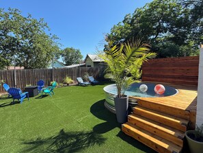 A backyard with artificial grass, a wooden deck, a hot tub, and blue chairs. - Gem On Pearl w/ Pool - Walk to Downtown Lockhart (Lockhart)