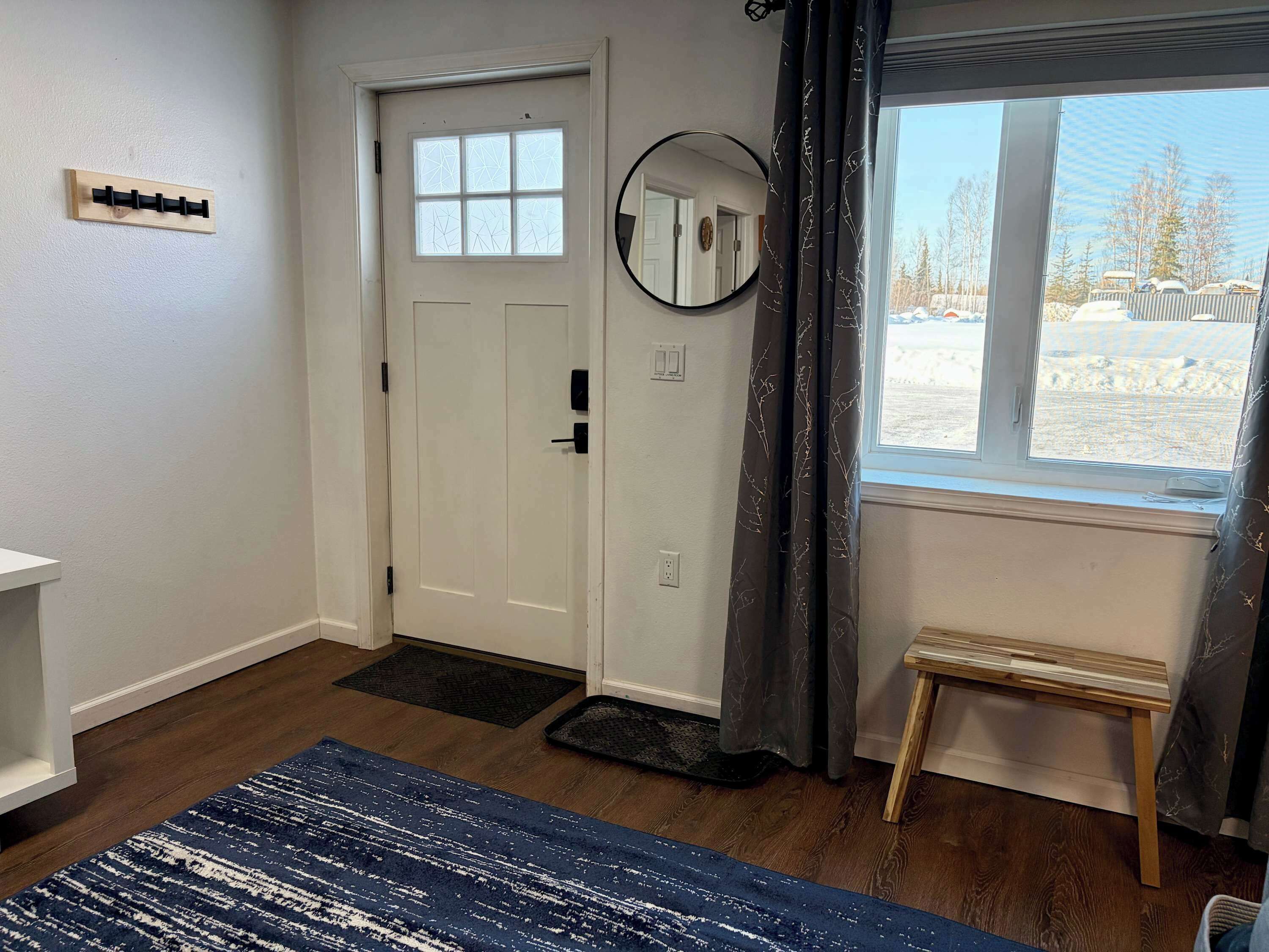 A room with a wooden floor, a white door with a window, a small wooden bench, a blue rug, and a mirror on the wall.