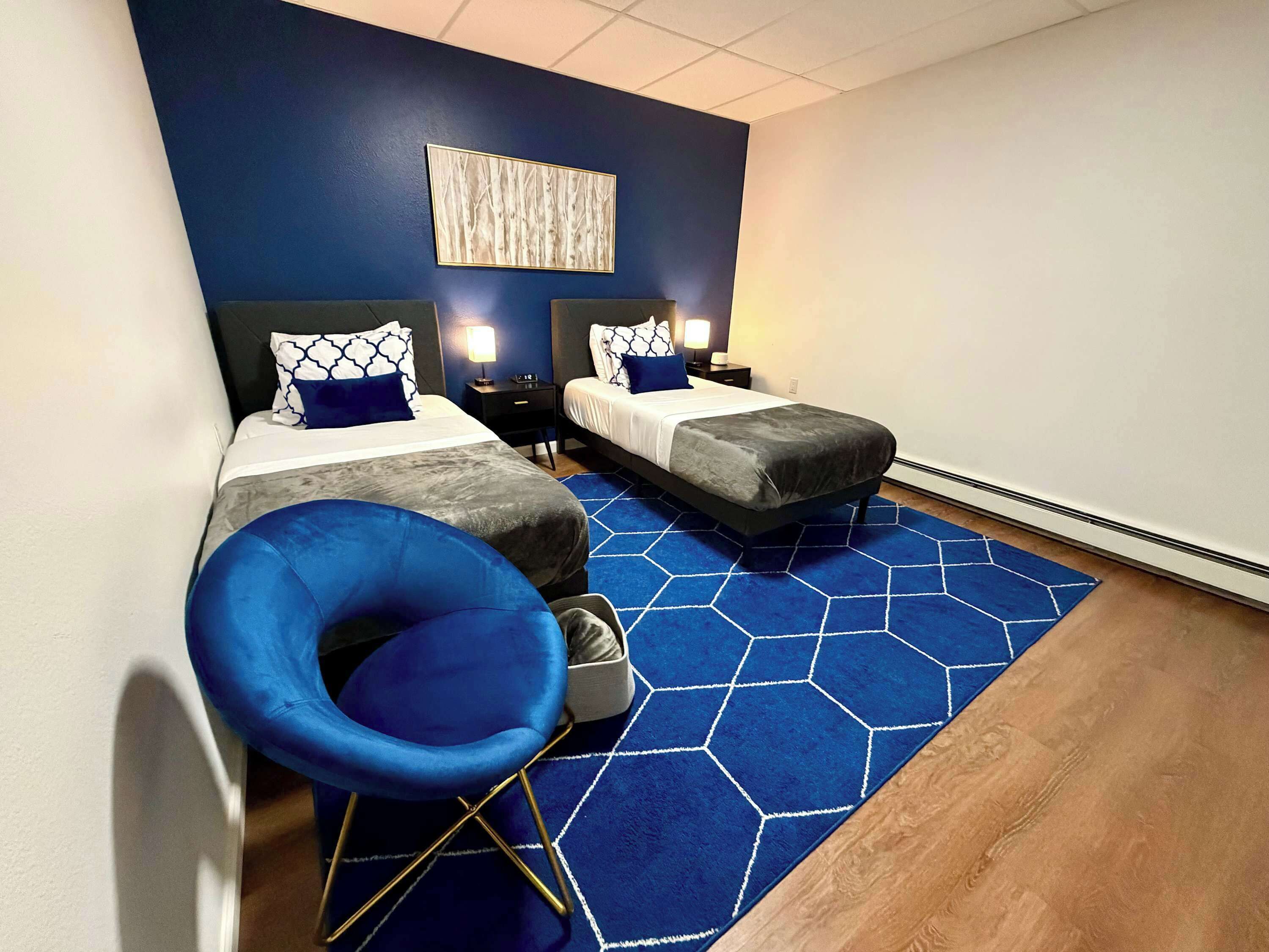 Une chambre d’hôtel avec deux lits, une chaise bleue, un tapis bleu en forme d’hexagone et une œuvre d’art murale.