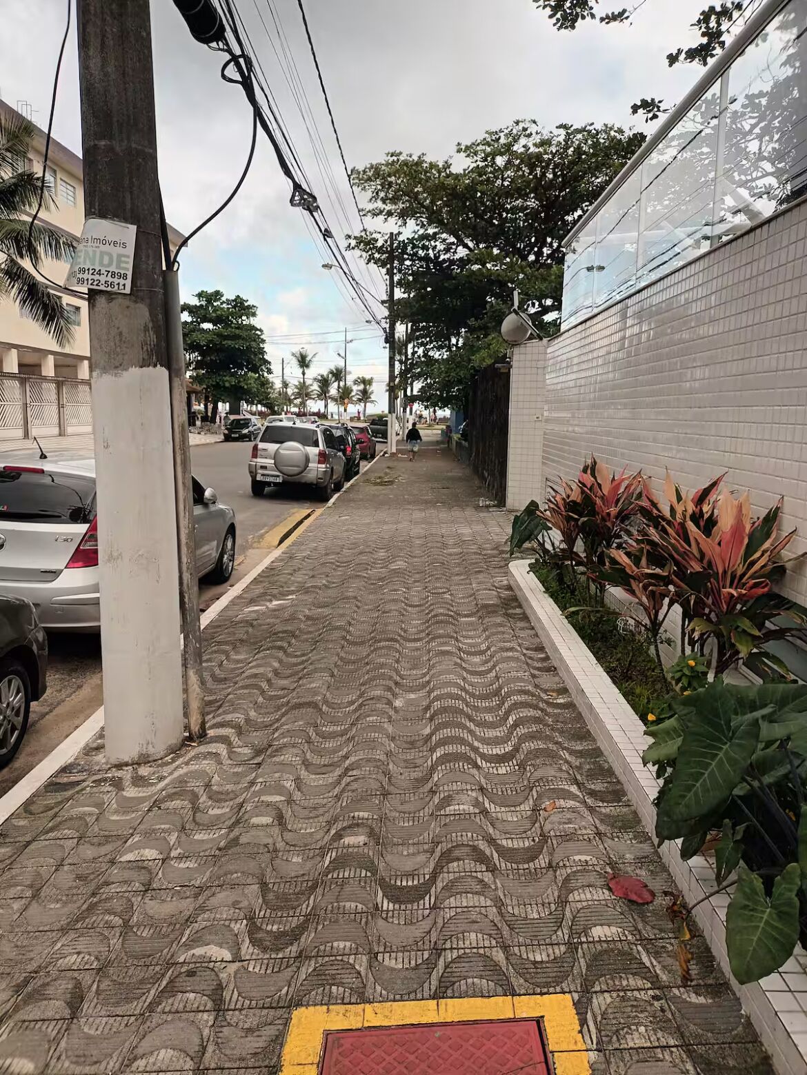 A sidewalk with patterned paving, parked cars, and a white building with a tiled wall.