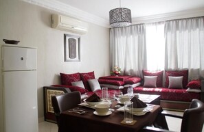 A dining area with a table set for four, a sofa with red and white cushions, a hanging light fixture, and a white refrigerator. - Isni appart hotel (Agadir)